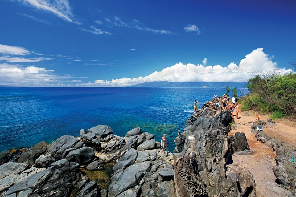 Visitors on the lava rocks of the Kapalua Coastal Trail above deep blue water, Molokaʻi in the distance